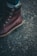 A close-up of a brown leather boot with metal eyelets and sturdy laces on a rough asphalt surface. The boot has thick, rugged soles, and a small brand logo is embossed on the side.