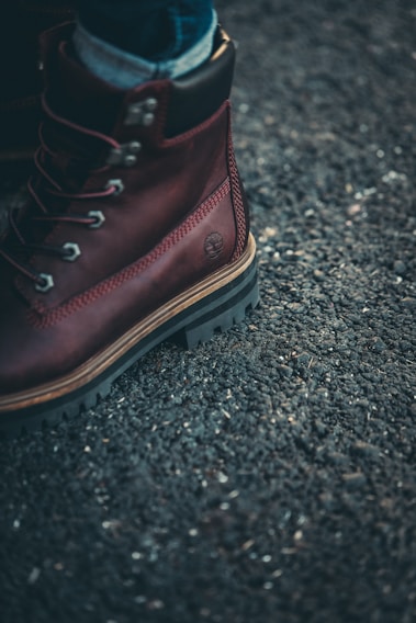 A close-up of a brown leather boot with metal eyelets and sturdy laces on a rough asphalt surface. The boot has thick, rugged soles, and a small brand logo is embossed on the side.