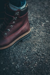 A close-up of a brown leather boot with metal eyelets and sturdy laces on a rough asphalt surface. The boot has thick, rugged soles, and a small brand logo is embossed on the side.