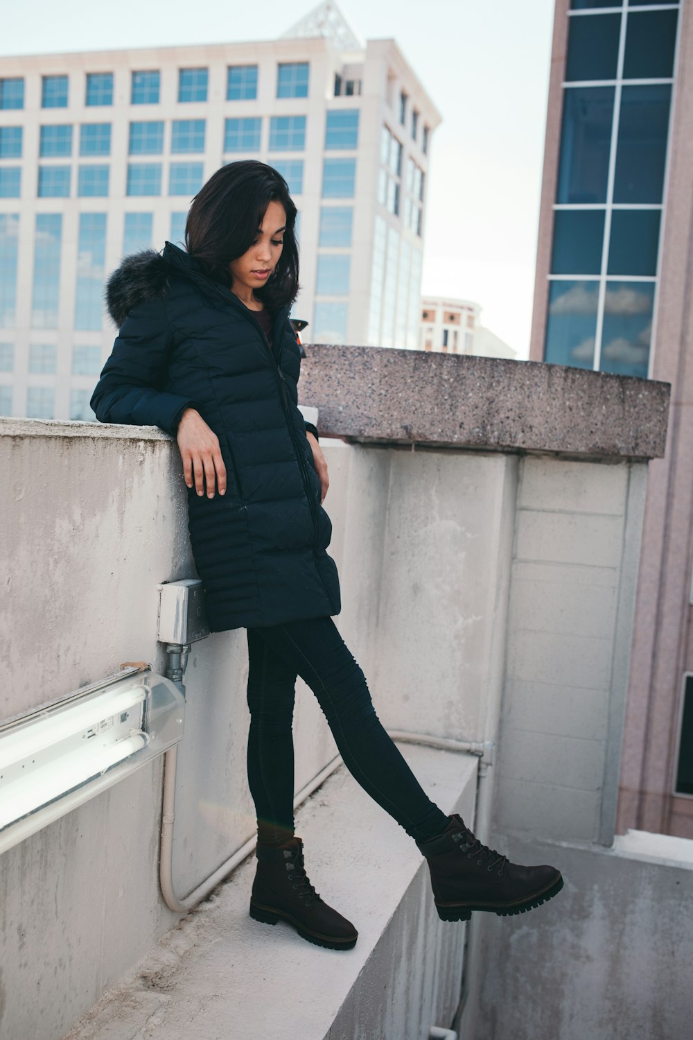 Frau trägt schwarzen Parka-Mantel, während sie sich an eine graue Betonwand lehnt