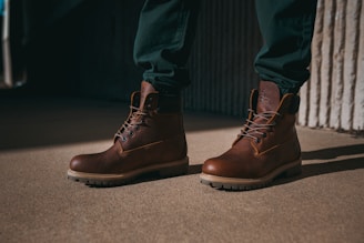 A pair of brown leather boots with thick soles is worn by a person standing on a sandy beige surface. The boots are laced up neatly, and the person is wearing dark green pants. Shadows cast on the ground suggest strong lighting, creating a contrast between the lit and shaded areas.