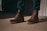 A pair of brown leather boots with thick soles is worn by a person standing on a sandy beige surface. The boots are laced up neatly, and the person is wearing dark green pants. Shadows cast on the ground suggest strong lighting, creating a contrast between the lit and shaded areas.