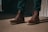A pair of brown leather boots with thick soles is worn by a person standing on a sandy beige surface. The boots are laced up neatly, and the person is wearing dark green pants. Shadows cast on the ground suggest strong lighting, creating a contrast between the lit and shaded areas.