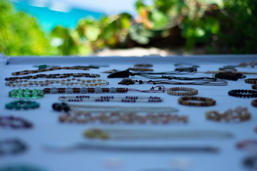 Various handmade bracelets and necklaces displayed on a white surface, with a blurred background of green foliage and a glimpse of blue sky. The jewelry pieces are made of colorful beads, shells, and stones, arranged neatly in rows.
