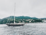 A large sailboat with a tall mast is floating on calm waters. In the background, there is a scenic view of a hillside covered in greenery and numerous small buildings or houses, resembling a coastal town. The sky appears overcast, creating a serene and tranquil atmosphere.