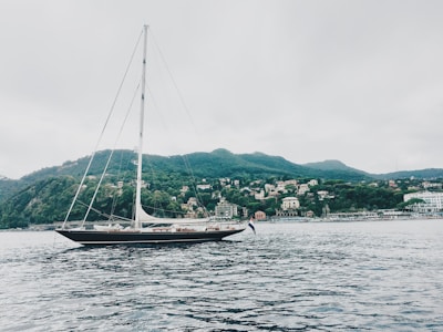 A large sailboat with a tall mast is floating on calm waters. In the background, there is a scenic view of a hillside covered in greenery and numerous small buildings or houses, resembling a coastal town. The sky appears overcast, creating a serene and tranquil atmosphere.