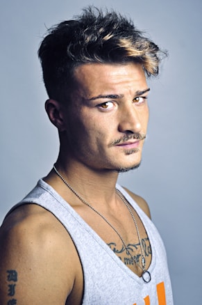 A young man with styled, wavy hair partially highlighted, wearing a gray tank top. He has a mustache and a short beard, along with visible tattoos on his upper chest and arm. He wears a chain necklace with a ring pendant and looks directly at the camera, set against a plain light background.