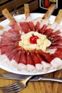 A white plate displays a visually appealing presentation of cured meat slices arranged in a circular pattern. In the center, there are shavings of cheese topped with a small cherry tomato. Breadsticks are strategically placed around the plate. A fork and knife are placed on the wooden table surface nearby.
