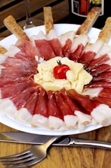 A white plate displays a visually appealing presentation of cured meat slices arranged in a circular pattern. In the center, there are shavings of cheese topped with a small cherry tomato. Breadsticks are strategically placed around the plate. A fork and knife are placed on the wooden table surface nearby.