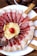 A plate artistically arranged with slices of cured meat, likely prosciutto, radiating in a circular pattern. At the center, there are shavings of hard cheese and a small red cherry tomato. Breadsticks are positioned decoratively around the plate. In the background, there is a bowl containing whole walnuts.