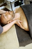 A person is lying on a spa bed with their eyes closed, receiving a neck massage. The individual is covered with a dark gray towel, and their hands are placed gently on their chest. The room is softly lit, and spa supplies are visible in the background.