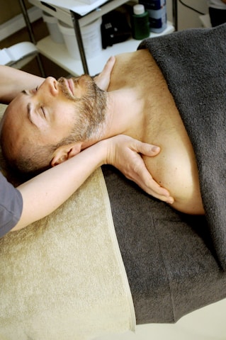 A person is lying on a spa bed with their eyes closed, receiving a neck massage. The individual is covered with a dark gray towel, and their hands are placed gently on their chest. The room is softly lit, and spa supplies are visible in the background.