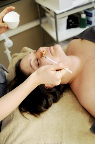 A person is lying on a treatment table in a spa-like setting, receiving a facial treatment. Another person is applying a facial mask with a brush while holding a small bowl. The space is neatly organized with shelves and skincare products in the background.