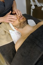 A person is lying on a massage table, covered with a dark towel, receiving a face massage. The massage therapist's hands are gently pressing on the person's forehead and eyes. The environment appears calm and relaxing.