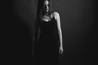 A monochrome photograph featuring a woman standing against a dark background. Her long hair partially obscures her face and she is wearing a simple, dark dress. The image is atmospheric and moody.