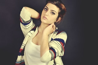 A woman poses with one arm raised, wearing a patterned cardigan over a white top. The background is dark, which contrasts with her light skin and the vibrant colors of the cardigan.