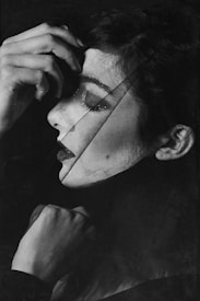 A black and white portrait capturing an artistic view of a woman's face in profile. The image is stylized with a textured overlay, creating a layered and abstract effect. Her hand is gently resting on her forehead, and her expression appears contemplative.