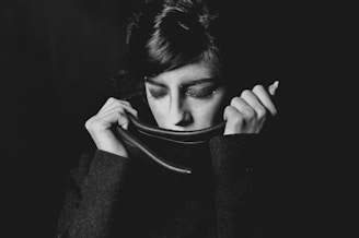 A person with closed eyes holds a high-collared jacket up to their face, creating a sense of introspection or contemplation. The image is in black and white, emphasizing shadows and highlights.