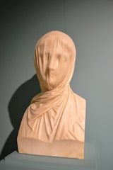 A marble bust of a veiled woman, intricately carved to depict fine details in the fabric draping over her face and shoulders. The sculpture captures the delicate folds of the veil, showcasing the artist's skill in rendering transparent and flowing materials in stone. The bust stands on a simple plinth, highlighted against a plain, muted background that draws attention to the craftsmanship.