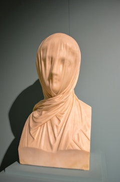 A marble bust of a veiled woman, intricately carved to depict fine details in the fabric draping over her face and shoulders. The sculpture captures the delicate folds of the veil, showcasing the artist's skill in rendering transparent and flowing materials in stone. The bust stands on a simple plinth, highlighted against a plain, muted background that draws attention to the craftsmanship.