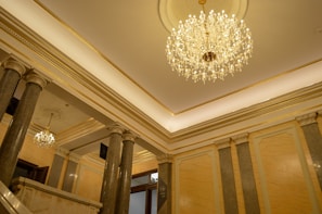 An opulent interior featuring a grand chandelier hanging from a high ceiling, complemented by columns and ornate architectural details. The walls and ceiling are adorned with luxurious gold accents and patterns, enhancing the overall elegance of the space.