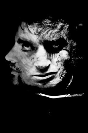 A monochromatic image featuring two overlapping faces creating a surreal and artistic composition. The faces are detailed with strong contrasts and shadows, producing an enigmatic appearance. The artistic lighting adds depth and texture to the overlapping features.