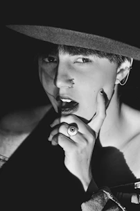 A person is captured in a black and white photograph wearing a dark wide-brimmed hat. They have piercings on their nose and ear, and a distinctive ring on their finger. The lighting creates strong contrasts, highlighting their facial features and casting shadows.