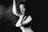 A black and white portrait of a person with a contemplative and abstract expression. The subject's face and body are partially illuminated, creating strong contrasts with the surrounding darkness. Their arm is raised, while their other hand rests thoughtfully on their shoulder, adding to the dramatic and artistic composition.