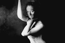 A black and white portrait of a person with a contemplative and abstract expression. The subject's face and body are partially illuminated, creating strong contrasts with the surrounding darkness. Their arm is raised, while their other hand rests thoughtfully on their shoulder, adding to the dramatic and artistic composition.