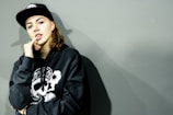 A person wearing a black hoodie with a graphic design on the front and a black cap looks at the camera with a relaxed expression. The individual has one hand raised to their mouth, partly covering it with their fingers. The background is a plain, muted gray color, creating a minimalistic and urban aesthetic.
