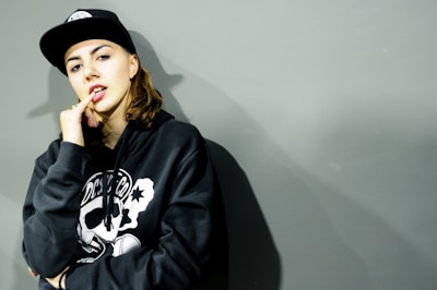A person wearing a black hoodie with a graphic design on the front and a black cap looks at the camera with a relaxed expression. The individual has one hand raised to their mouth, partly covering it with their fingers. The background is a plain, muted gray color, creating a minimalistic and urban aesthetic.