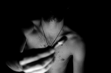 A black and white photograph of a shirtless person holding a matchstick towards the camera. The lighting highlights the contours of the body while the background remains dark, creating a dramatic contrast.
