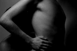 The image shows a close-up of two bare torsos intertwined, with one hand gently placed on the waist of the other person. The lighting is dim, creating deep shadows that highlight the forms and contours of the bodies.