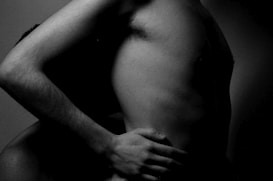 The image shows a close-up of two bare torsos intertwined, with one hand gently placed on the waist of the other person. The lighting is dim, creating deep shadows that highlight the forms and contours of the bodies.