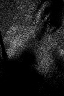 A textured dark surface with indistinct shapes and shadows creating a mysterious atmosphere. It appears as if there are faint facial features emerging from the shadows, enhancing the enigmatic feel.