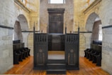 A historic interior with stone walls and wooden pews in rows along the sides. A dark wooden bimah with ornate carvings sits at the center, surrounded by arches and stonework, suggesting a religious setting.