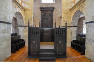 A historic interior with stone walls and wooden pews in rows along the sides. A dark wooden bimah with ornate carvings sits at the center, surrounded by arches and stonework, suggesting a religious setting.