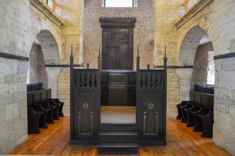 A historic interior with stone walls and wooden pews in rows along the sides. A dark wooden bimah with ornate carvings sits at the center, surrounded by arches and stonework, suggesting a religious setting.