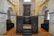 A historic interior with stone walls and wooden pews in rows along the sides. A dark wooden bimah with ornate carvings sits at the center, surrounded by arches and stonework, suggesting a religious setting.