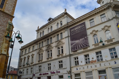A historic multi-story building with classical architectural features, including ornate window frames and decorative elements. The facade displays a large poster advertising the 'Srebrenica Exhibition'. Street lamps with a vintage design are visible in the foreground, and part of an adjacent building can be seen on the left side.