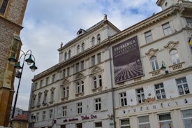 A historic multi-story building with classical architectural features, including ornate window frames and decorative elements. The facade displays a large poster advertising the 'Srebrenica Exhibition'. Street lamps with a vintage design are visible in the foreground, and part of an adjacent building can be seen on the left side.