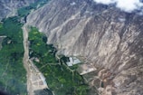 Aerial view of a landscape featuring vast green agricultural fields bordered by rugged, rocky mountain slopes. A small settlement and some scattered buildings, including what appears to be a greenhouse or industrial structure, are visible near the base of the mountains. The contrast between the lush greenery and the barren mountains creates a striking visual effect.