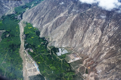 Aerial view of a landscape featuring vast green agricultural fields bordered by rugged, rocky mountain slopes. A small settlement and some scattered buildings, including what appears to be a greenhouse or industrial structure, are visible near the base of the mountains. The contrast between the lush greenery and the barren mountains creates a striking visual effect.