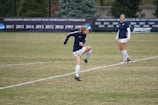 A soccer player is mid-action on a grassy field, wearing a navy-blue jersey and white shorts. Another player in the same uniform is standing nearby, observing. In the background, a black banner displays various years of tournament appearances, surrounded by evergreen trees.