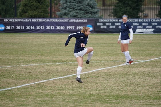 A soccer player is mid-action on a grassy field, wearing a navy-blue jersey and white shorts. Another player in the same uniform is standing nearby, observing. In the background, a black banner displays various years of tournament appearances, surrounded by evergreen trees.