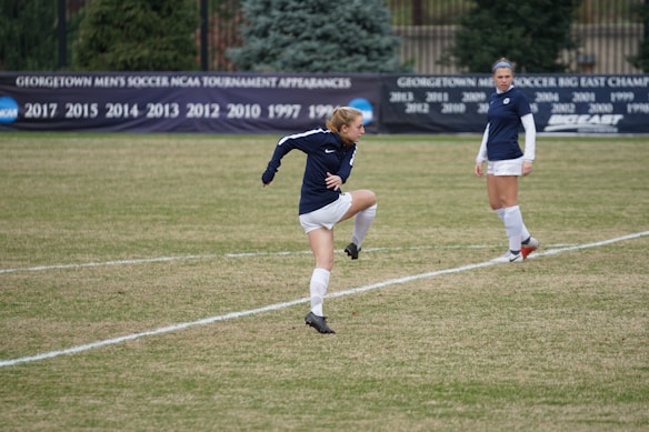 A soccer player is mid-action on a grassy field, wearing a navy-blue jersey and white shorts. Another player in the same uniform is standing nearby, observing. In the background, a black banner displays various years of tournament appearances, surrounded by evergreen trees.