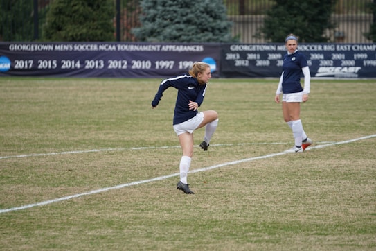 A soccer player is mid-action on a grassy field, wearing a navy-blue jersey and white shorts. Another player in the same uniform is standing nearby, observing. In the background, a black banner displays various years of tournament appearances, surrounded by evergreen trees.