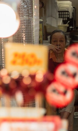 An elderly person is sitting indoors smoking a cigarette. The foreground is blurred with bright lights and signs, creating a bokeh effect. The background shows kitchen items and furniture, giving a sense of being in a cozy, cluttered space.