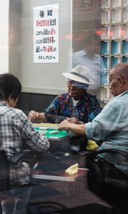 A group of elderly men are engaged in a game of mahjong inside a room. One man is wearing a white hat and colorful shirt, while others wear casual shirts. Mahjong tiles are spread out on the green table. A sign with Japanese text is visible on the wall, and stacks of tiles are organized nearby. Reflections on the glass give the scene a candid, intimate feel.