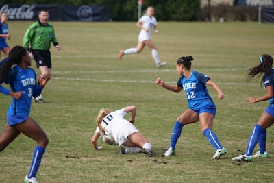 A soccer match featuring female players in blue and white uniforms. One player in a white uniform appears to have fallen near the ball, while other players in blue are positioned around her. A referee wearing a green jersey is in the background observing the game. The scene takes place on a grass field.